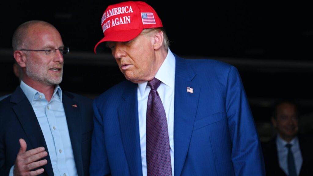 FCC Commissioner Brendan Carr standing next to and speaking to Donald Trump, who is wearing a "Make America Great Again" hat.