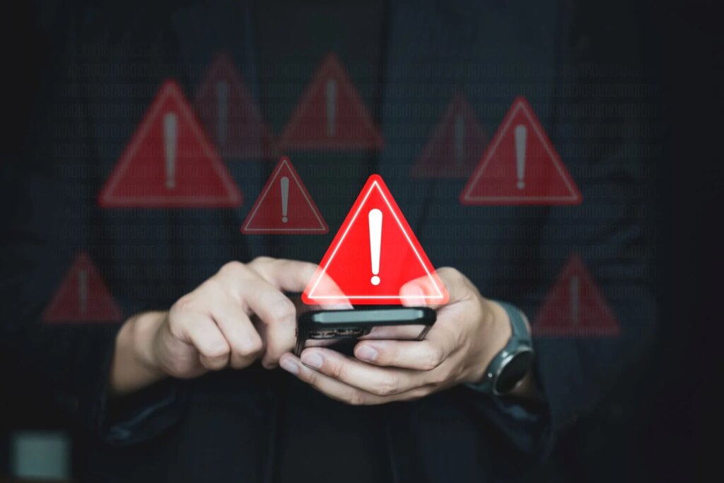 A pair of hands holding a phone underneath a red triangle with a white exclamation mark in it, signifying a warning.