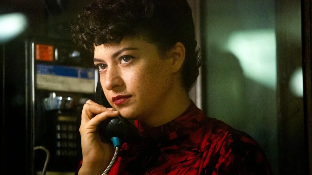 Production still from Search Party showing Alia Shawkat wearing a red dress on the phone in a phone booth.