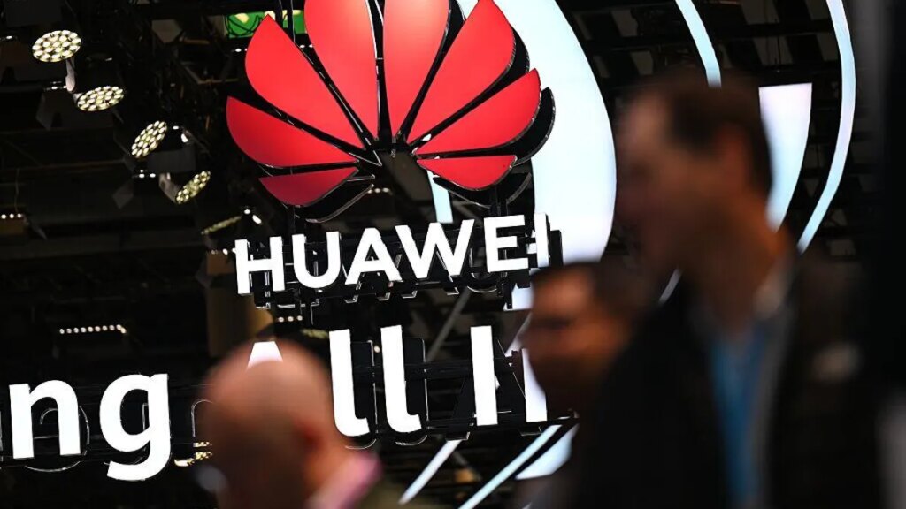 Huawei's logo hangs from the ceiling as several blurry people pass by the camera in the foreground.
